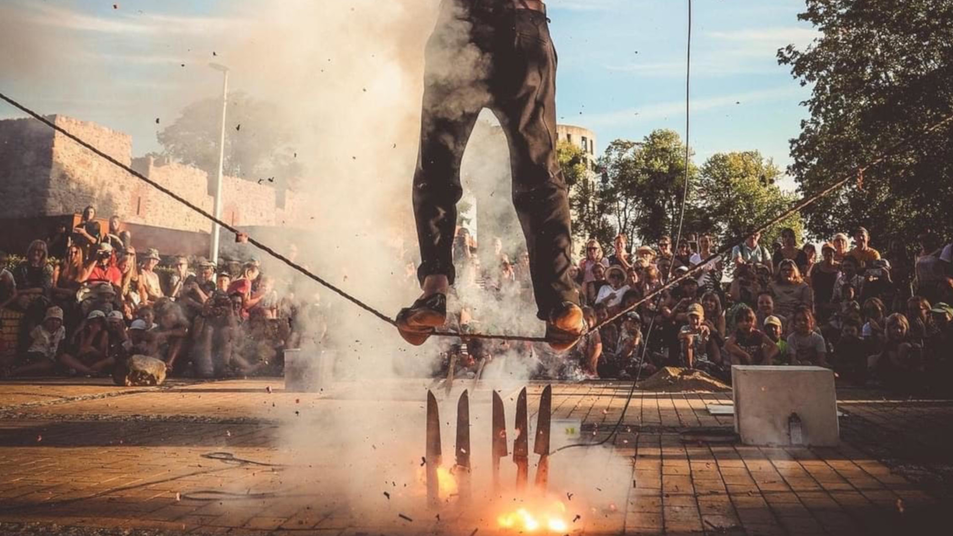 Mężczyzna chodzi po taśmie. Pod stopami ma ustawione pionowo noże i ogień.
